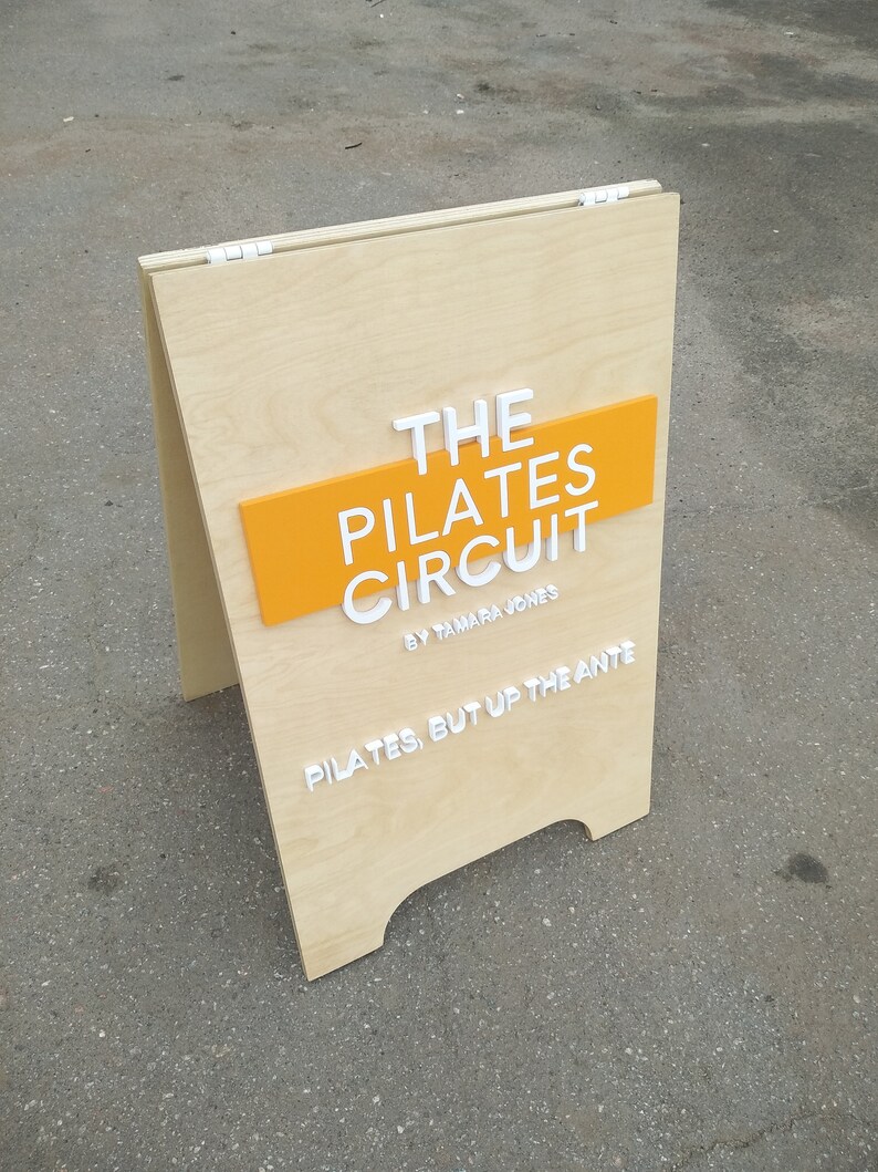 K&ouml;nnte beinhalten: Ein A-f&ouml;rmiges Schild aus Holz mit dem Text "THE PILATES CIRCUIT BY TAMARA JONES" in wei&szlig;en Buchstaben auf gelbem Hintergrund. Das Schild tr&auml;gt auch den Text "PILATES, BUT UP THE ANTE" in wei&szlig;en Buchstaben unten.