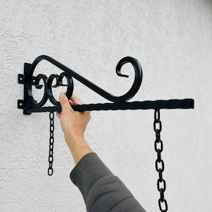 Könnte beinhalten: Schwarzer Metall-Schildhalter mit einem dekorativen Schnörkeldesign und zwei Ketten. Der Halter ist an einer weißen Wand montiert.