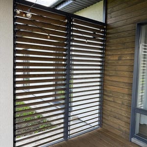 May include: A wooden slat fence with a dark metal frame. The fence is made of horizontal wooden slats, creating a slatted design. String lights are strung above the fence. The fence is on a wooden deck.
