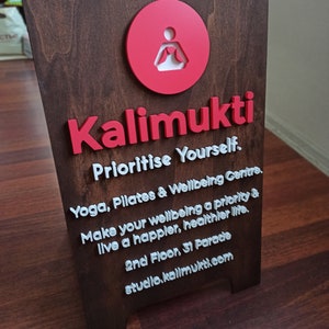 K&ouml;nnte beinhalten: Ein braunes Holzschild mit wei&szlig;er Beschriftung, auf dem steht "Kalimukti Prioritise Yourself. Yoga, Pilates & Wellbeing Centre. Make your wellbeing a priority & live a happier, healthier life. 2nd Floor, 31 Parade studio.kalimukti.com". Das Schild zeigt auch einen roten Kreis mit einer wei&szlig;en Silhouette einer Person in einer Yoga-Pose.