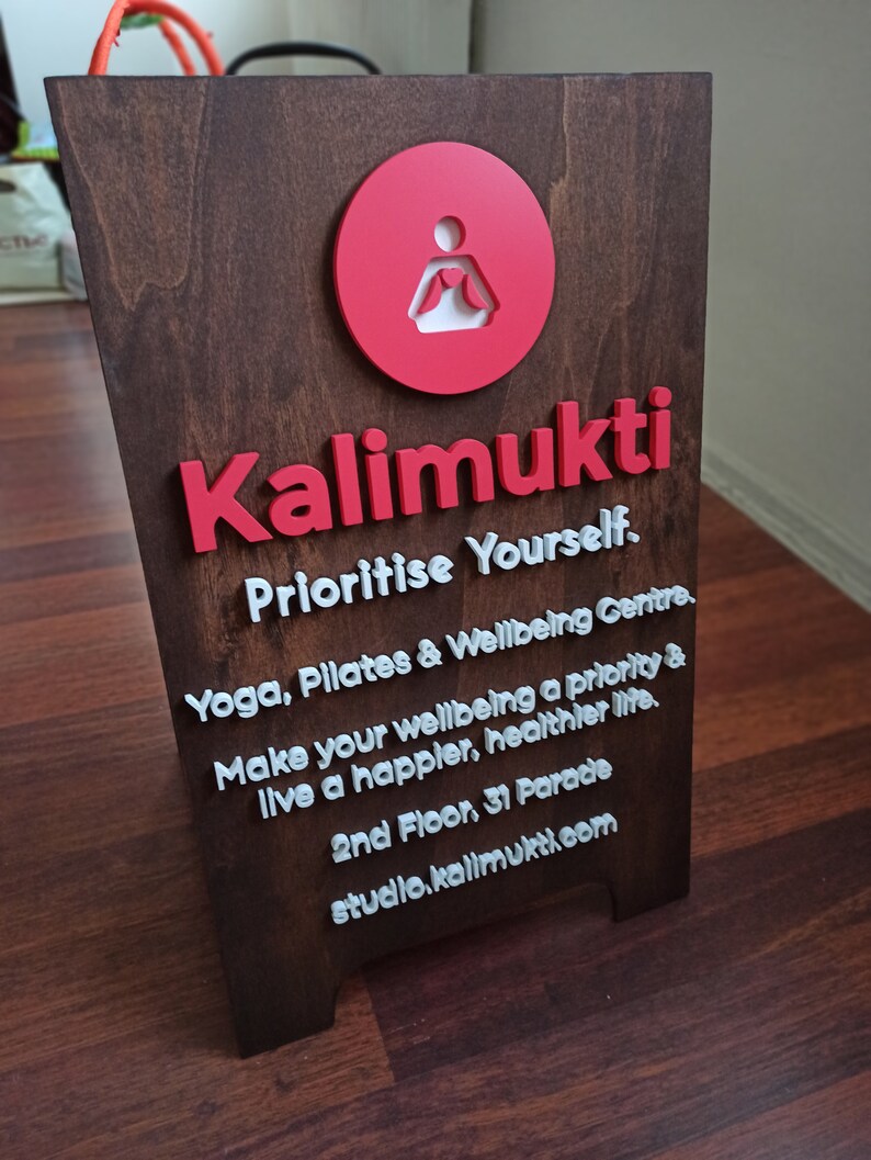K&ouml;nnte beinhalten: Ein braunes Holzschild mit wei&szlig;er Beschriftung, auf dem steht "Kalimukti Prioritise Yourself. Yoga, Pilates & Wellbeing Centre. Make your wellbeing a priority & live a happier, healthier life. 2nd Floor, 31 Parade studio.kalimukti.com". Das Schild zeigt auch einen roten Kreis mit einer wei&szlig;en Silhouette einer Person in einer Yoga-Pose.
