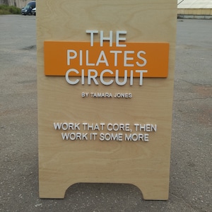 K&ouml;nnte beinhalten: Ein Schild aus Holz mit orangefarbenem Hintergrund und wei&szlig;em Text, der "THE PILATES CIRCUIT BY TAMARA JONES" lautet. Das Schild hat auch wei&szlig;en Text, der "WORK THAT CORE, THEN WORK IT SOME MORE" lautet.