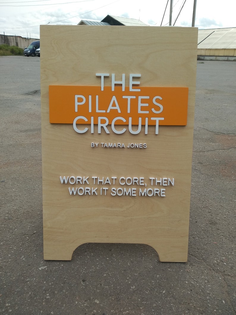 K&ouml;nnte beinhalten: Ein Schild aus Holz mit orangefarbenem Hintergrund und wei&szlig;em Text, der "THE PILATES CIRCUIT BY TAMARA JONES" lautet. Das Schild hat auch wei&szlig;en Text, der "WORK THAT CORE, THEN WORK IT SOME MORE" lautet.