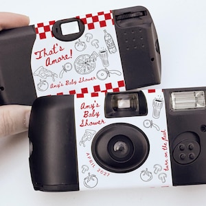 May include: Two black disposable cameras with custom labels. One camera reads "That's Amore! Amy's Baby Shower" with pizza and drink illustrations. The other camera reads "Amy's Baby Shower April 2027". The phrase "Camera Not Included" is at the bottom.