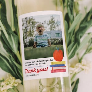 May include: A clear wine bottle with a custom label featuring a photo of a child and the text "Thank you!" with an apple and book graphic. The bottle is surrounded by white flowers and green foliage.