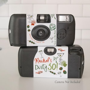 May include: Two black disposable cameras stacked on top of each other. Each camera has a white label with cocktail illustrations and the text "Rachel's Dirty 30!". The phrase "Camera Not Included" is printed at the bottom.