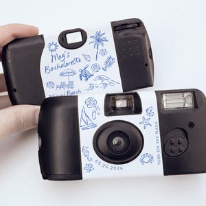 May include: Two black disposable cameras with white labels featuring blue illustrations of beach elements and text. One camera reads "Meg's Bachelorette Miami Beach 05.25.2026". The other camera has the text "TURN ON THE FLASH".