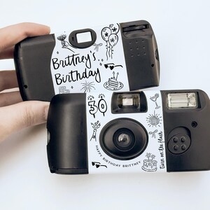 May include: Two black disposable cameras with white decorative labels. The labels feature the text "Brittney's Birthday" and "Happy Birthday Brittney" with doodle-style illustrations of balloons, cake, and fireworks. The text "Turn on the flash" is also visible.