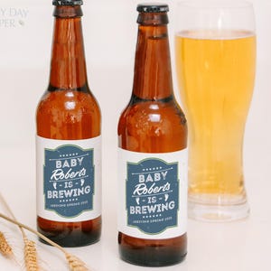 May include: Two amber glass beer bottles with white labels featuring a blue rectangular border and white text that reads Baby Roberts is Brewing Arriving Spring 2025. The bottles have black caps and are positioned next to a glass of golden beer.