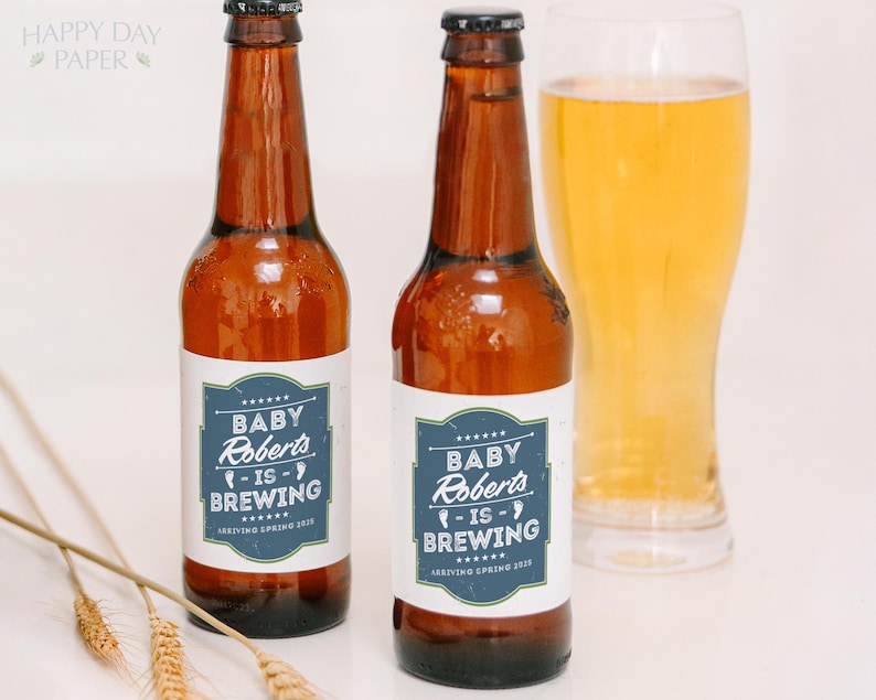 May include: Two amber glass beer bottles with white labels featuring a blue rectangular border and white text that reads Baby Roberts is Brewing Arriving Spring 2025. The bottles have black caps and are positioned next to a glass of golden beer.