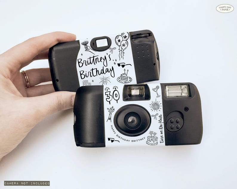 May include: Two black disposable cameras with white labels featuring the text "Brittney's Birthday" and decorative doodles. One camera displays the number "30". The phrase "Turn on the flash" is also visible.