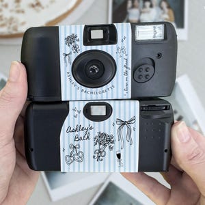 May include: Two black disposable cameras wrapped in custom light blue and white striped paper labels. The labels feature black line art illustrations of bouquets, bows, and champagne glasses, with the text Ashley's Bachelorette and turn on the flash.