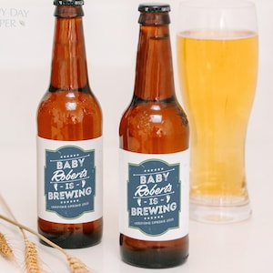 May include: Two amber glass beer bottles with white labels featuring a blue rectangular border and white text that reads Baby Roberts is Brewing Arriving Spring 2025. The bottles have black caps and are positioned next to a glass of golden beer.