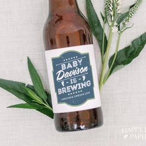 May include: A brown glass bottle featuring a white rectangular label with a blue border. The label displays the text BABY Davison IS BREWING and ARRIVING SPRING 2025, accented by small white footprints and stars, accompanied by green leaves.