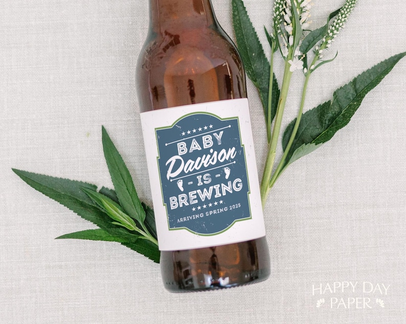 May include: A brown glass bottle featuring a white rectangular label with a blue border. The label displays the text BABY Davison IS BREWING and ARRIVING SPRING 2025, accented by small white footprints and stars, accompanied by green leaves.