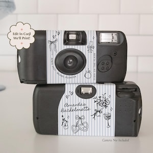 May include: Two black disposable cameras wrapped in light blue and white striped paper labels. The top label features a ring, bow, and text reading April 2022 Miami, Florida. The bottom label features a bouquet, glass, and Amanda's Bachelorette.