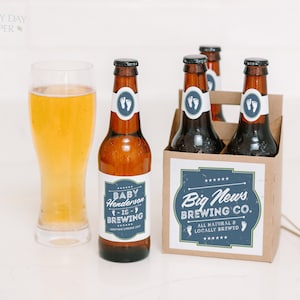 May include: A glass of golden beer next to brown glass bottles in a cardboard carrier. The labels feature blue and white graphics with text reading Baby Henderson is Brewing, Big News Brewing Co., and All Natural & Locally Brewed with baby feet.