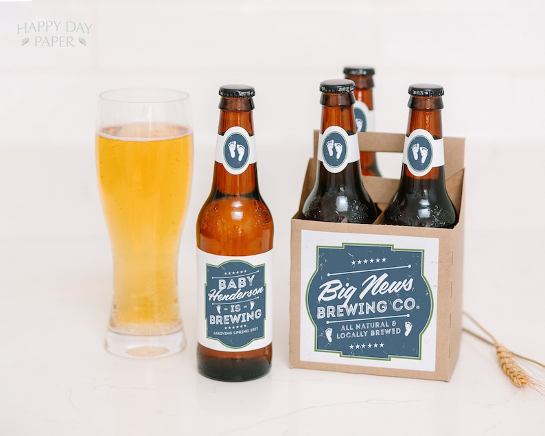 May include: A glass of golden beer next to brown glass bottles in a cardboard carrier. The labels feature blue and white graphics with text reading Baby Henderson is Brewing, Big News Brewing Co., and All Natural & Locally Brewed with baby feet.