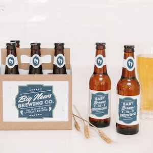 May include: A brown cardboard six-pack carrier and two amber glass beer bottles with white and blue labels. The labels feature the text Big News Brewing Co. All Natural & Locally Brewed and Baby Browns Is Brewing with baby footprint icons.