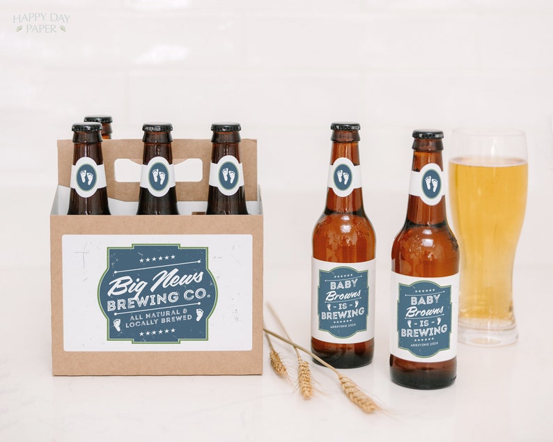 May include: A brown cardboard six-pack carrier and two amber glass beer bottles with white and blue labels. The labels feature the text Big News Brewing Co. All Natural & Locally Brewed and Baby Browns Is Brewing with baby footprint icons.