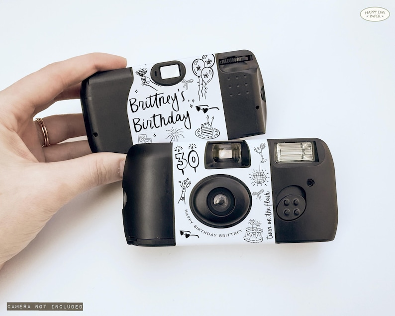 May include: Two black disposable cameras with white decorative labels. The labels feature the text "Brittney's Birthday" and "Happy Birthday Brittney" with doodle-style illustrations of balloons, cake, and fireworks. The text "Turn on the flash" is also visible.