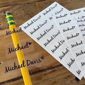 May include: Sheets of clear labels printed with the name "Michael" and "Michael Davis" in a cursive font. A yellow pencil with a pink eraser is in the foreground. The labels are for organizing and labeling items.