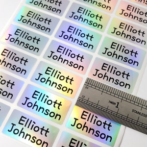 May include: A sheet of rectangular holographic stickers, each printed with the name Elliott Johnson in black sans-serif font. A stainless steel ruler is placed across the stickers, showing each individual label is approximately 1 inch wide.