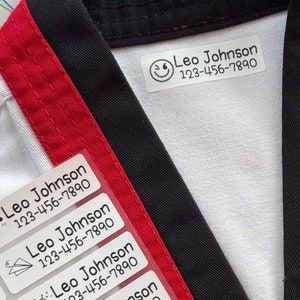 May include: White fabric with a black and red stripe. Several white rectangular labels with black text and icons are attached to the fabric. The labels read "Leo Johnson 123-456-7890". A wooden ruler with inch markings is visible in the bottom right corner.