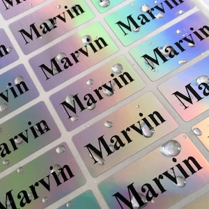 May include: A sheet of rectangular adhesive labels with a holographic, iridescent finish. Each label features the name Marvin printed in a bold, black serif font. Several clear water droplets are scattered across the surface of the labels.