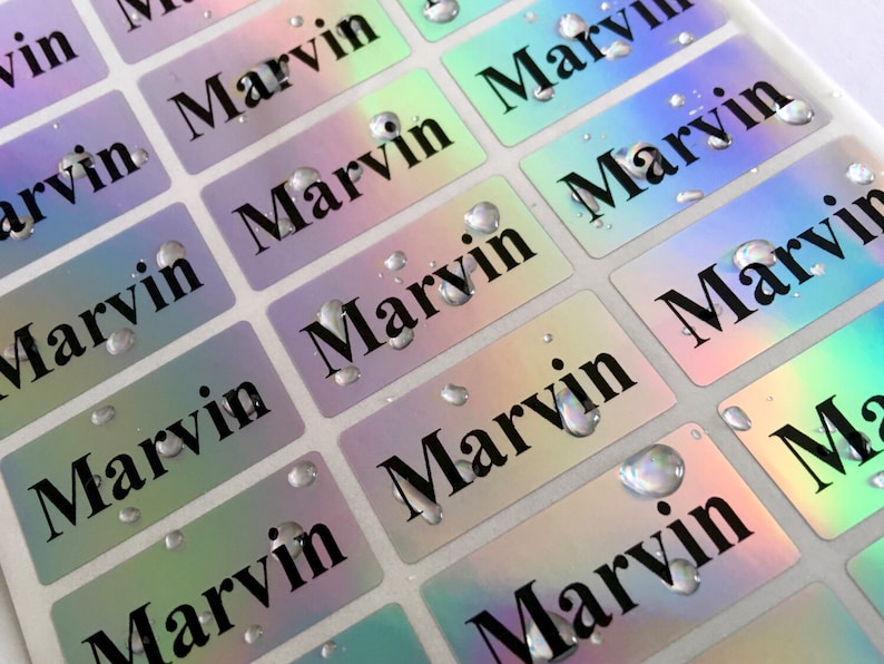 May include: A sheet of rectangular adhesive labels with a holographic, iridescent finish. Each label features the name Marvin printed in a bold, black serif font. Several clear water droplets are scattered across the surface of the labels.