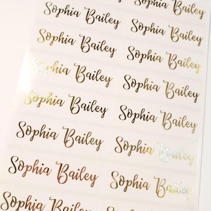 May include: Sheet of gold foil name labels. Each label reads "Sophia Bailey" in a cursive font. The labels are arranged in rows and columns on a white background. The foil has a shiny, reflective quality.