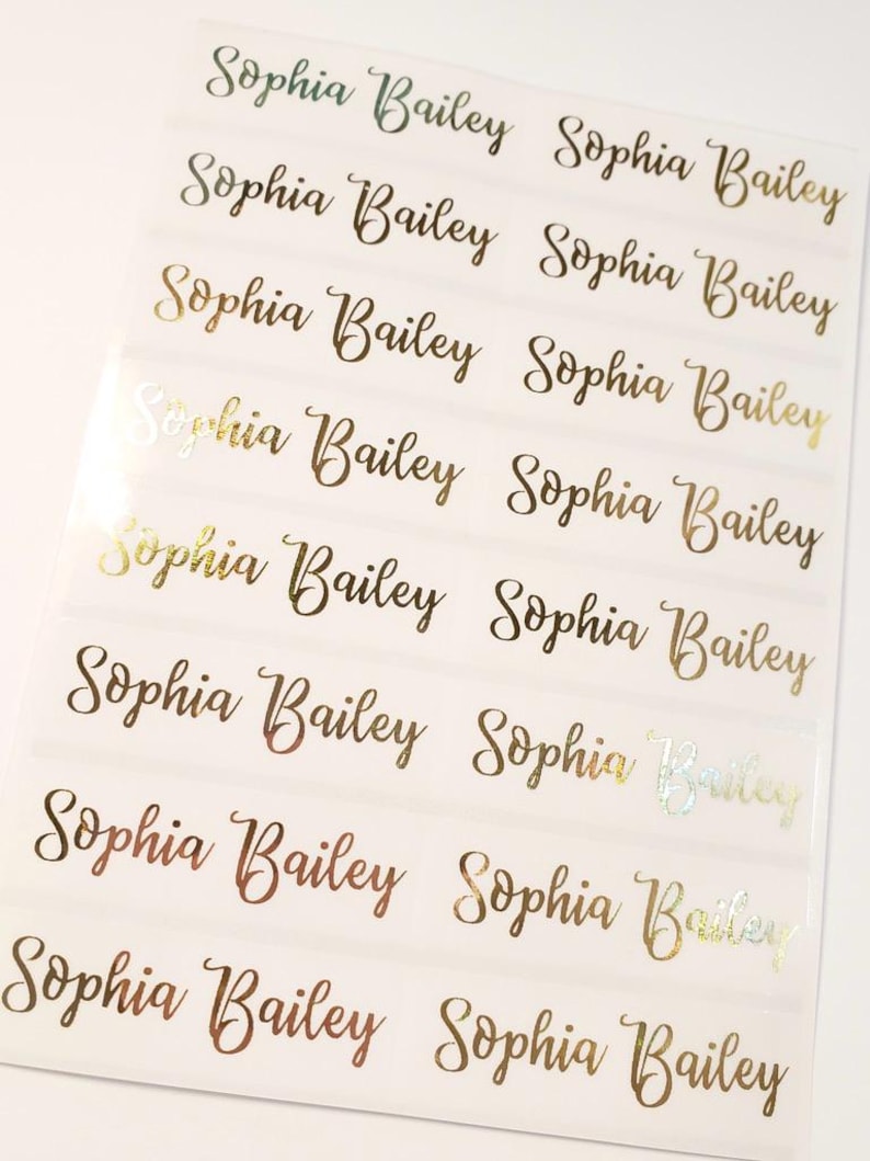 May include: Sheet of gold foil name labels. Each label reads "Sophia Bailey" in a cursive font. The labels are arranged in rows and columns on a white background. The foil has a shiny, reflective quality.