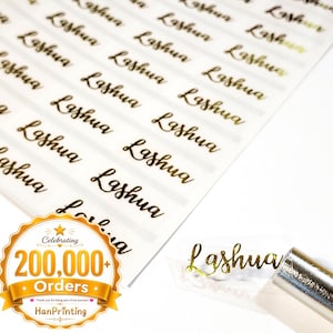 Puede incluir: Una hoja de etiquetas adhesivas transparentes con el nombre Lashua impreso en una fuente cursiva de color dorado metálico. Una etiqueta es levantada por una herramienta de metal, revelando el acabado dorado reflectante sobre el soporte transparente.