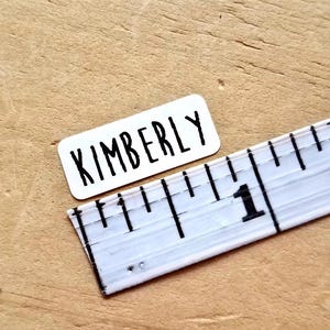 May include: A white rectangular sticker with the name "KIMBERLY" in black, bold letters. A white ruler with black markings and the number "1" is next to the sticker. The objects are on a light brown wooden surface.