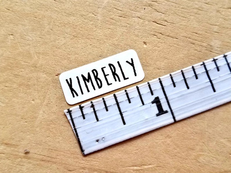 May include: A white rectangular sticker with the name "KIMBERLY" in black, bold letters. A white ruler with black markings and the number "1" is next to the sticker. The objects are on a light brown wooden surface.