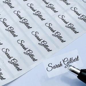 May include: A sheet of rectangular, transparent adhesive labels featuring the name Sarah Gilbert printed in a black, cursive script font. One individual label is being peeled away from the backing sheet using the tip of a black pen.
