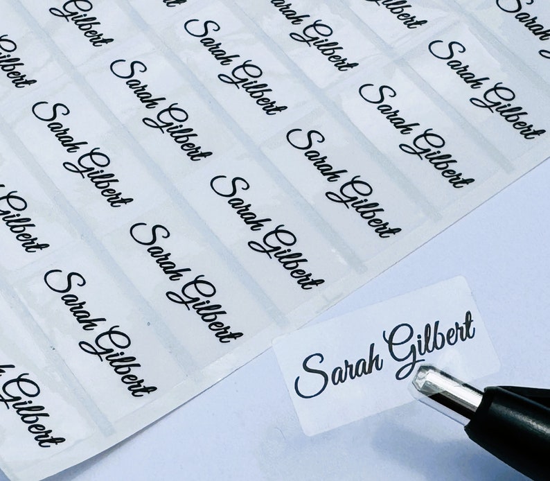 May include: A sheet of rectangular, transparent adhesive labels featuring the name Sarah Gilbert printed in a black, cursive script font. One individual label is being peeled away from the backing sheet using the tip of a black pen.