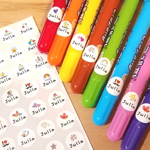 以下が含まれることがあります： 「Julia」という名前と、ハート、星、虹などのさまざまなデザインが施されたカラフルな丸いステッカーのシート。ステッカーは、カラフルなマーカーのセットの隣にあります。