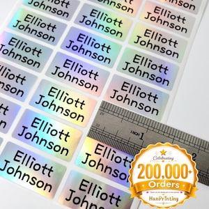 May include: A sheet of rectangular holographic stickers, each printed with the name Elliott Johnson in black sans-serif font. A metal ruler is placed over the stickers, showing each individual label measures approximately 1 inch in width.