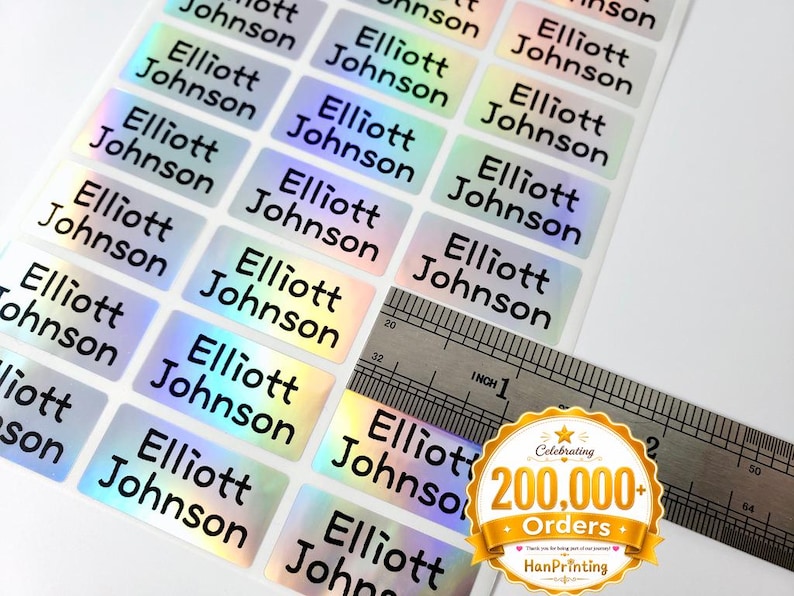 May include: A sheet of rectangular holographic stickers, each printed with the name Elliott Johnson in black sans-serif font. A metal ruler is placed over the stickers, showing each individual label measures approximately 1 inch in width.