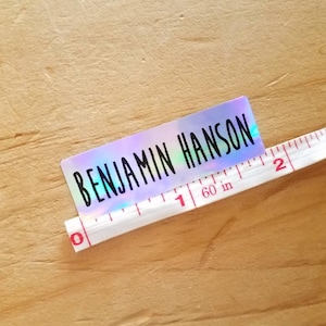 May include: A holographic sticker with the name "BENJAMIN HANSON" in black lettering. A white measuring tape is placed next to the sticker, showing measurements in inches. The sticker is approximately 2 inches long.