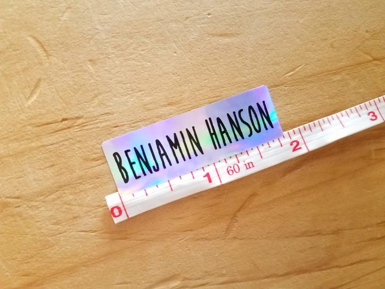 May include: A holographic sticker with the name "BENJAMIN HANSON" in black lettering. A white measuring tape is placed next to the sticker, showing measurements in inches. The sticker is approximately 2 inches long.