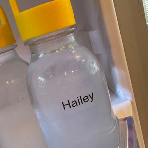May include: A clear glass bottle with a bright yellow plastic cap featuring a handle. The bottle contains a cloudy white liquid and has a white rectangular label on the front with the name Hailey printed in black sans-serif font.