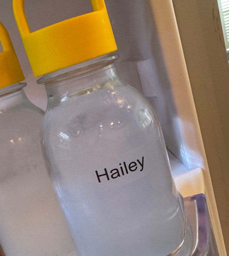 May include: A clear glass bottle with a bright yellow plastic cap featuring a handle. The bottle contains a cloudy white liquid and has a white rectangular label on the front with the name Hailey printed in black sans-serif font.
