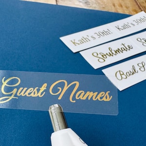 May include: A collection of labels with gold lettering on a blue background. The labels include the words "Guest Names", "Kath's 30th!", "Soulmate", and "Basil Salt". A white tool is positioned near the "Guest Names" label.