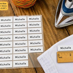 May include: A sheet of white rectangular iron-on clothing labels with the name Michelle printed in black sans-serif font. Each label measures 1.18 inches by 0.59 inches. One label is placed on white fabric next to a wooden ruler for scale.