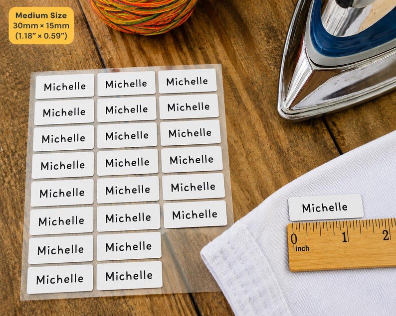 May include: A sheet of white rectangular iron-on clothing labels with the name Michelle printed in black sans-serif font. Each label measures 1.18 inches by 0.59 inches. One label is placed on white fabric next to a wooden ruler for scale.