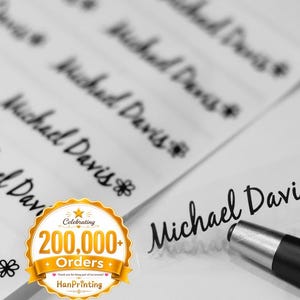 May include: A sheet of clear adhesive labels featuring the black printed name Michael Davis followed by a small flower icon. A black pen rests near one detached label. A gold circular badge displays the text Celebrating 200,000+ Orders HanPrinting.