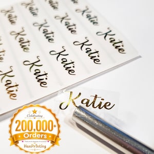 May include: A sheet of clear adhesive labels featuring the name Katie in a gold metallic cursive script. One individual label is detached and positioned near a silver metal tool. A gold circular badge displays the text Celebrating 200,000+ Orders.