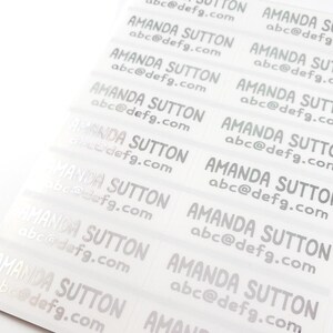 May include: A sheet of clear labels printed with the name "AMANDA SUTTON" and the email address "abc@defg.com" in a silver font. The labels are arranged in rows and columns, ready for use.